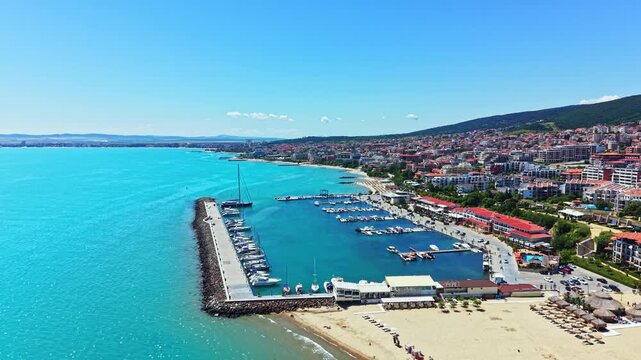 View of a marina with several boats docked in blue water next to a sandy beach. Town with houses and cafes is visible in the background under a bright sky. People may be enjoying the area.