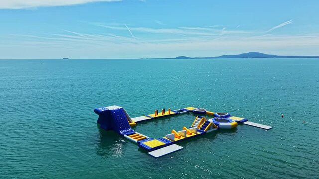 Children and adults enjoy a water activity park in the sea. Bright blue and yellow inflatable structures float on the water under a clear sky. Waves gently move around the park.