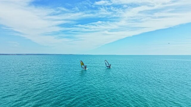 Two people enjoy windsurfing on clear blue waters under a bright sky. The sun shines, making for a lovely afternoon at the beach. Waves are gentle as they navigate their boards.