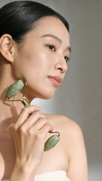 Vertical video: Starting skincare Asian woman rolling jade roller up jaw in studio bare shoulders