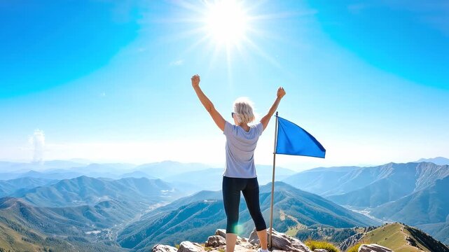Blonde woman celebrating achievement on mountain peak with blue flag. Panning shot of hiker with raised arms overlooking vast valley.