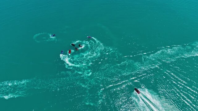 Several individuals enjoy riding jet skis on the blue water close to the shore. The sun shines down while waves create patterns in the sea.