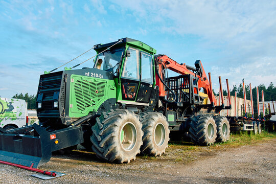 A green Noe NF210-8R forestry forwarder with a crane arm and a log trailer parked on a gravel lot under a blue sky.