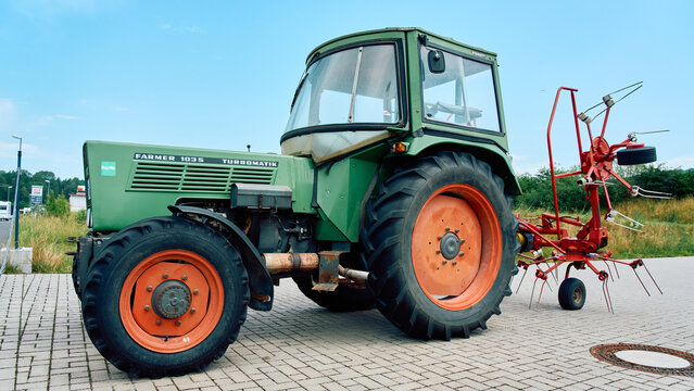 Side view of a vintage green Fendt Farmer 103S tractor with a red hay tedder attachment, parked on a paved lot on a sunny day.
