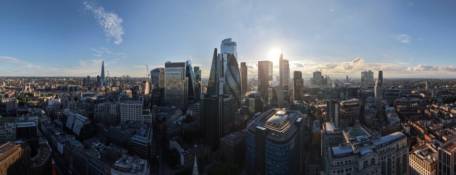 Aerial view of the City of London skyline featuring iconic skyscrapers like The Gherkin and The Shard under a bright sun, with dense urban architecture in London, England, United Kingdom.
