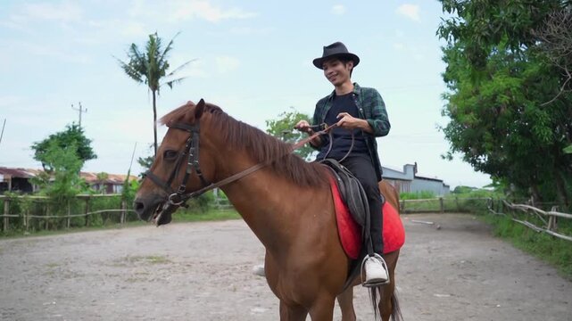 Full clip follows rider on brown horse moving calmly across gravel yard in rural setting, with trees, fence, simple houses, and soft daylight shifting from