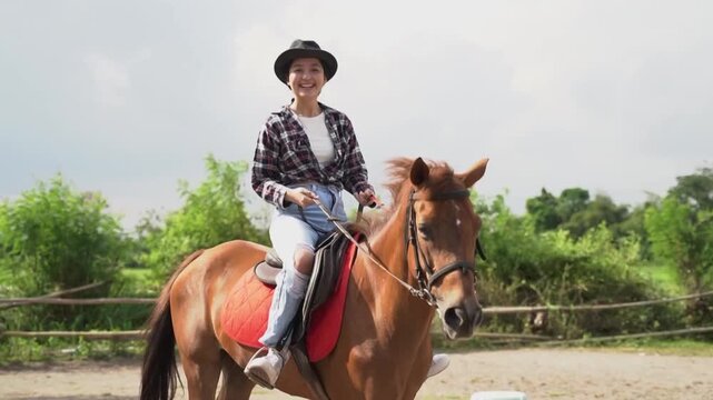 Near-end frame shows smiling woman riding brown horse near center of frame, moving slowly across sandy paddock beside wooden fence and green trees under