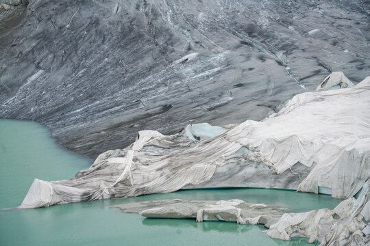 Aerial view of the Rhone Glacier covered with white protective blankets to prevent melting, situated next to a turquoise glacial lake in Obergoms, Valais, Switzerland.