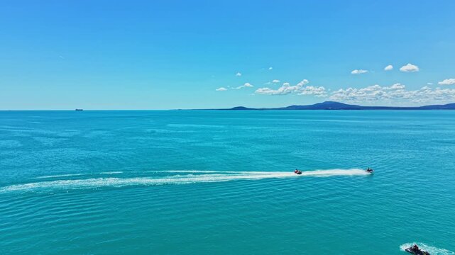 Two people ride jet skis across calm blue water. The sun shines bright in the sky. Waves form behind the jet skis as they speed along the coast. A distant landform is visible.