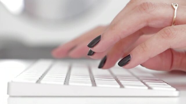 Close-up of a woman's hands with black manicure typing on a white keyboard, Female fingers with an elegant ring typing text on a computer, Detail shot of a woman working at her office desk typing fast