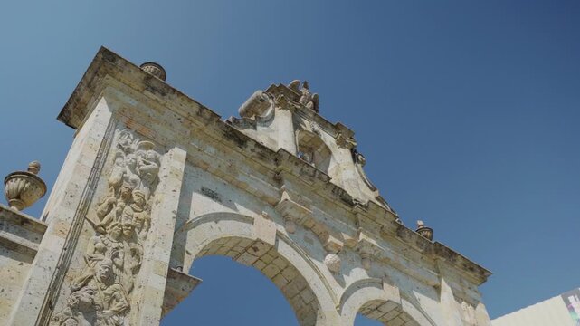 Aerial drone shot looking up at the Zapopan Arch, capturing its decorative columns and eagle sculpture at the top against a vivid blue sky in Guadalajara, Jalisco