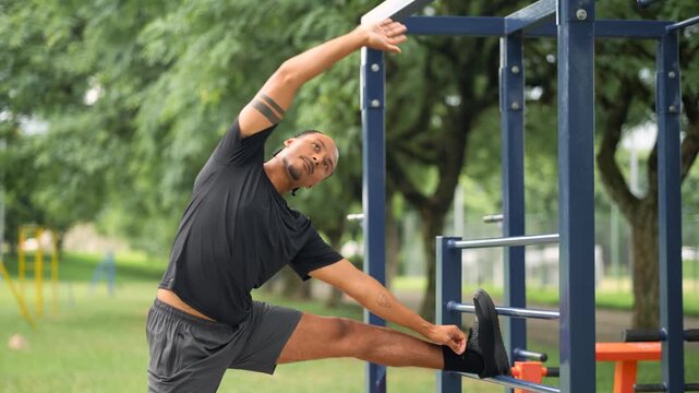 Young Black Latin man in sportswear doing side stretch using outdoor gym bar equipment in urban park during daytime