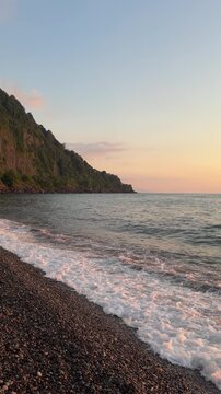 Gentle sea waves washing over a pebble beach during sunset. Peaceful coastal landscape with rocky shoreline and warm evening light. Vertical 4K footage.