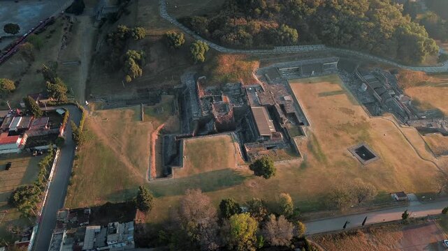 Aerial drone over San Andres Cholula archaeological zone at dawn, with warm orange sunrise light illuminating ancient pyramid ruins and a pre-Hispanic sacrifice area in Puebla, Mexico