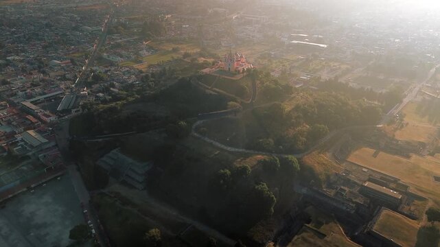 Aerial drone orbits the hilltop church atop the Cholula pyramid at dawn, with the Puebla skyline spreading below and the Popocatepetl and Iztaccihuatl volcanoes rising on the horizon