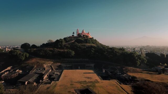 Aerial drone orbits the hilltop church atop the Cholula pyramid at dawn, with the Puebla skyline spreading below and the Popocatepetl and Iztaccihuatl volcanoes rising on the horizon