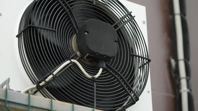 Close-up of an industrial HVAC condenser fan with protective metal grille and exposed electrical wiring on an exterior wall, showing mechanical details of the outdoor cooling unit.
