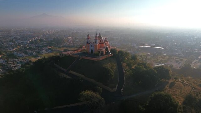 Aerial drone orbits the hilltop church atop the Cholula pyramid at dawn, with the Puebla skyline spreading below and the Popocatepetl and Iztaccihuatl volcanoes rising on the horizon