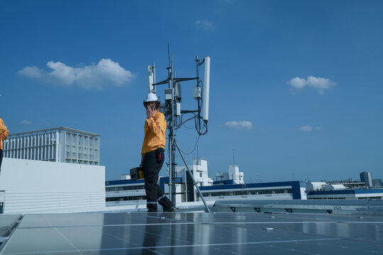 Technician pair in yellow safety jackets discuss work beside a rooftop telecom antenna on a rooftop solar farm, showing sustainable power, inspection and maintenance on a rooftop.