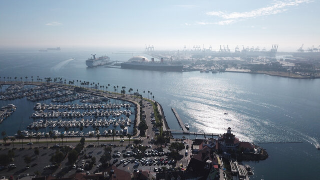 Aerial view of the Queen Mary docked in Long Beach harbor, with a marina filled with boats, palm trees, and the city skyline in the background, Long Beach, California, United States.