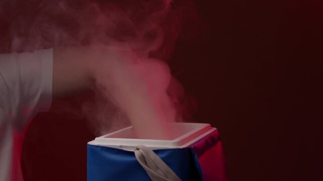 Courier placing frozen cold pack into portable thermal bag for transporting sensitive materials, with white vapor from dry ice billowing out against dark red background, close-up, slow motion