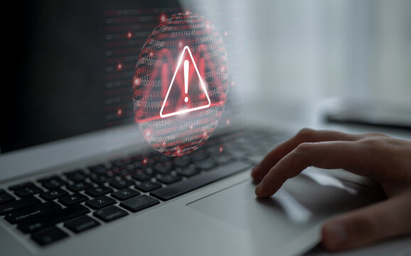 A close-up of a hand hovering over a laptop touchpad with a digital warning symbol and binary code in the background, illustrating cybersecurity threats and risk awareness. Gantry