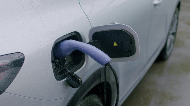 Electric vehicle charging process with a blue charging cable inserted into the charging port of a silver car parked in a wet environment with visible raindrops