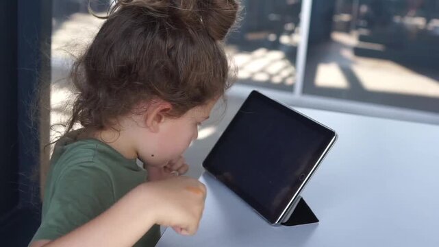 A young girl sitting at a table tapping, scrolling, and interacting with the screen of a tablet computer supported by a case stand.