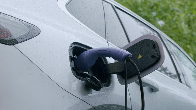 Electric vehicle charging process with charging cable inserted into port, rain droplets on car surface, and green foliage in the background enhancing the scene's natural ambiance