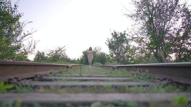 Woman walking on railway track towards camera in summer. Girl in yellow dress strolls along sunny railroad line. Female steps on wooden sleepers outdoors. Young person walks forward in nature.