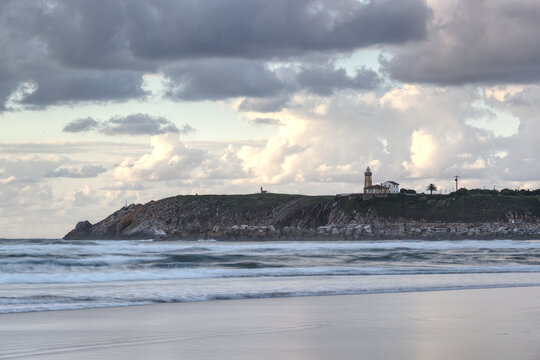 The majestic San Juan de Nieva lighthouse stands on a cliff at sunset, viewed from the sandy beach of Aviles, Asturias. Warm golden hour light.
