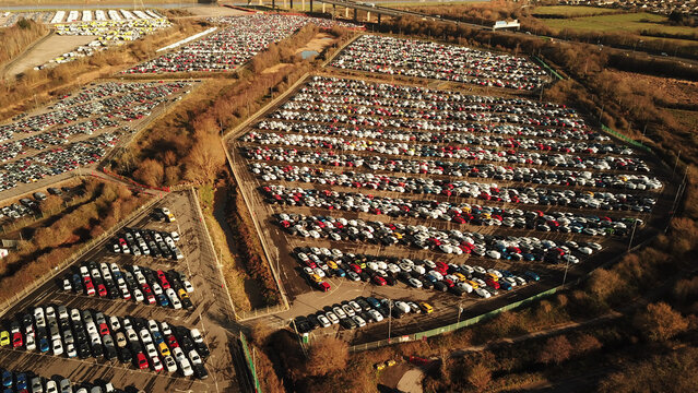 Aerial view of a large new car storage lot filled with rows of parked vehicles, surrounded by trees and a highway overpass in Gloucestershire, United Kingdom.