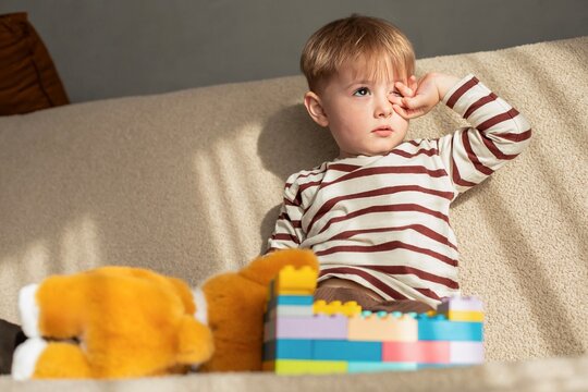 Tired little boy rubbing his eye while sitting on sofa with toys. Cute toddler boy in striped long sleeve shirt looking sleepy and rubbing irritated eye near colorful plastic building blocks.