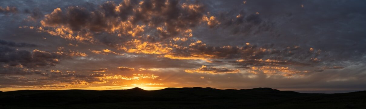 Panorama of spectacular sunset with bright orange and purple colors and thin clouds with the horizon as a silhouette of rolling hills. Light shafts extend from the sun
