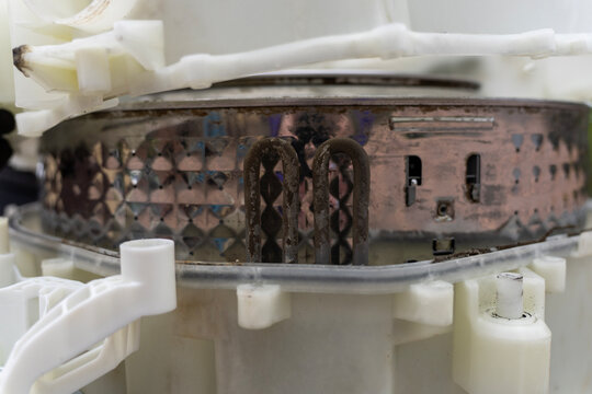Close up of a dirty limescale covered heating element installed inside a disassembled washing machine stainless steel drum during appliance repair service.