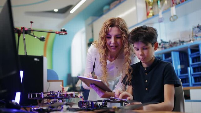 Tracking shot woman guiding boy assembling drone and programming electronics in stem classroom lateral pan.