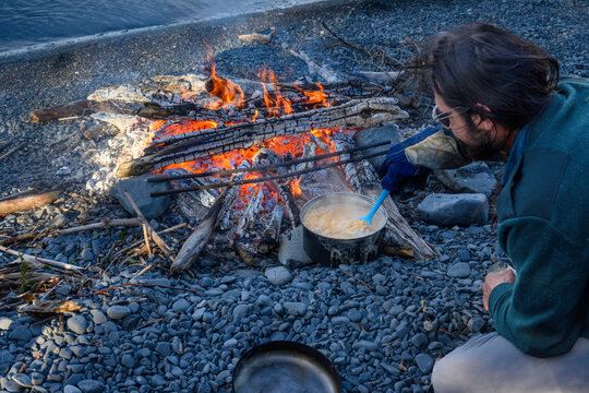 Guide cooking lentils over campfire at dusk | A sea kayak guide wearing a fire glove stirs a pot of lentils over a beach campfire at dusk, with the lake visible behind.