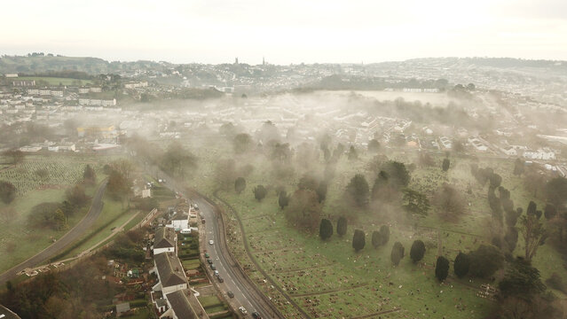 Aerial view of a foggy town with a cemetery, winding road, and residential houses in Torquay, England, United Kingdom.