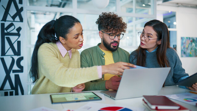 Wide view of corporate team planning campaign. Three colleagues studying printed charts. Male comparing laptop data. Woman holding tablet and smiling. Brainstorming marketing strategy in glass office.