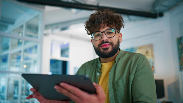 Corporate office scene with focused employee. Bearded male wearing glasses analyzing data on tablet. Tapping screen and reviewing statistics. Papers and water glass on desk. Managing finance tasks.