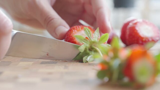 Hand cuts tops off fresh strawberries with knife on cutting board. Concept of fruit preparation and summer cooking