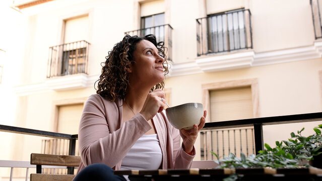 Woman with curly hair eating from a bowl on a city balcony, appreciating the morning scenery and relaxing