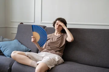Fototapeten Musik Woman at home suffering from overheating, sitting on sofa in living room holding hand paper fan for cooling down. Tired young female on couch with sick expression, overwhelmed by summer heat or fever  © DimaBerlin