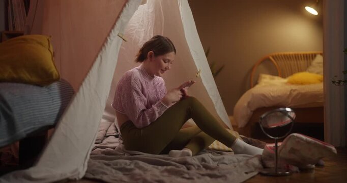 Young woman using a smartphone inside a cozy bedroom teepee at home