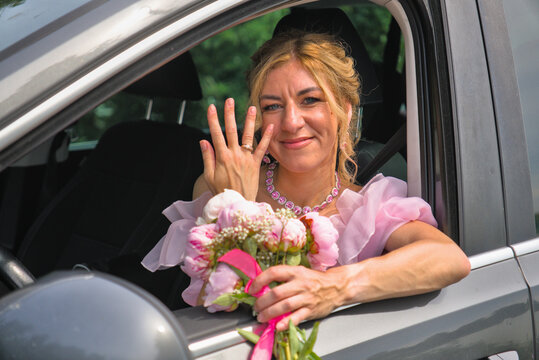 Joyful bride in a pink wedding gown smiling and showing her engagement ring from a car window.