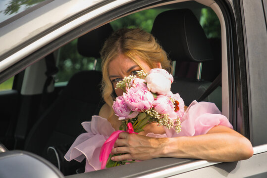 Beautiful bride in a luxury pink dress hiding her face behind a delicate bouquet of peonies while sitting in a wedding car.