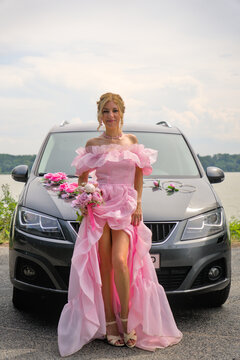 Attractive woman in a fluffy long pink dress with a high slit standing in front of a decorated wedding car.