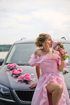 Sensual young woman in a fluffy pink dress sitting on a car hood and smelling a bouquet of peonies.