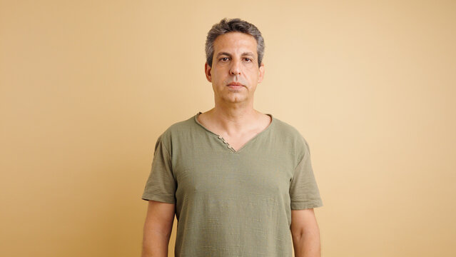 Serious middle aged man standing, looking directly at camera. Plain beige background, studio portrait with natural light