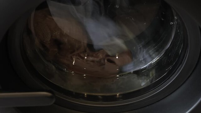 Close-up 4k video of industrial washer operating with a steady, moderate rotation, actively cleaning dirty garments inside a modern public laundry facility